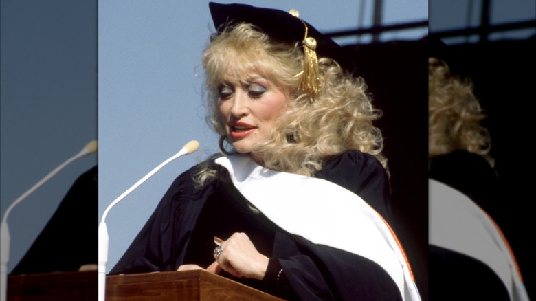 Dolly Parton speaks into a microphone while wearing a graduation gown.