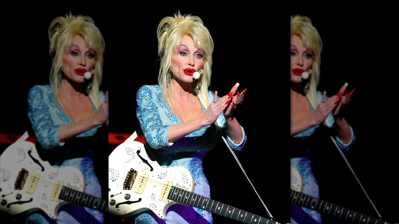 Dolly Parton, wearing a blue outfit, claps her hands while singing into a microphone.