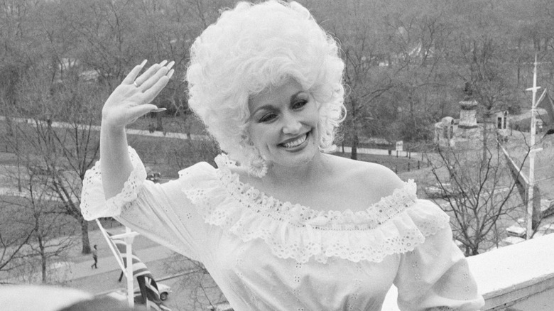 Dolly Parton, wearing a retro outfit with gathered details, waves while smiling.