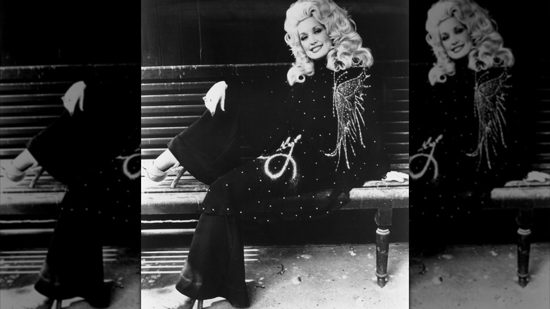 Dolly Parton poses with one leg resting on a bench and the other on the ground.