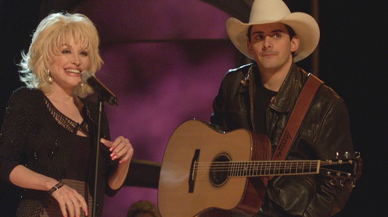 Dolly Parton laughs while standing behind a microphone as Bradley Paisely looks into the distance.