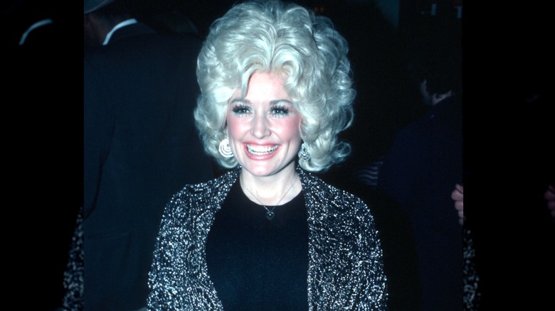 Dolly Parton, wearing a black outfit with a shimmery cover, flashes a broad smile.