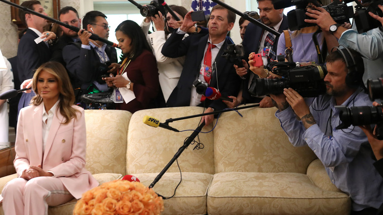 Melania Trump seated on sofa with swarm of reporters behind her