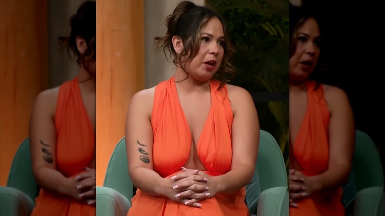 Liz Woods, wearing an orange outfit, speaks while her hands rest on her knees.