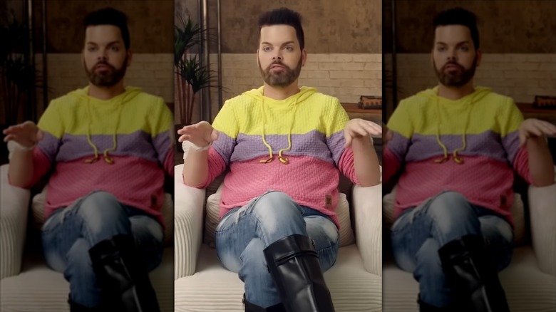 Tim Malcolm, wearing a colorful sweater and black boots, gesturing with his hands.