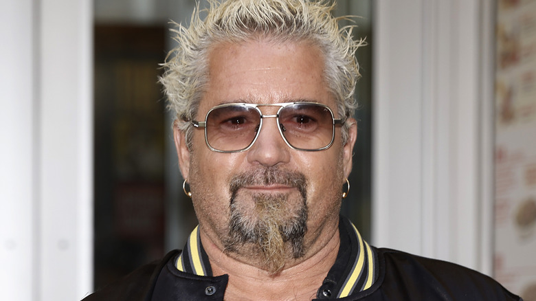 Guy Fieri posing for cameras in a black varsity jacket and tinted glasses