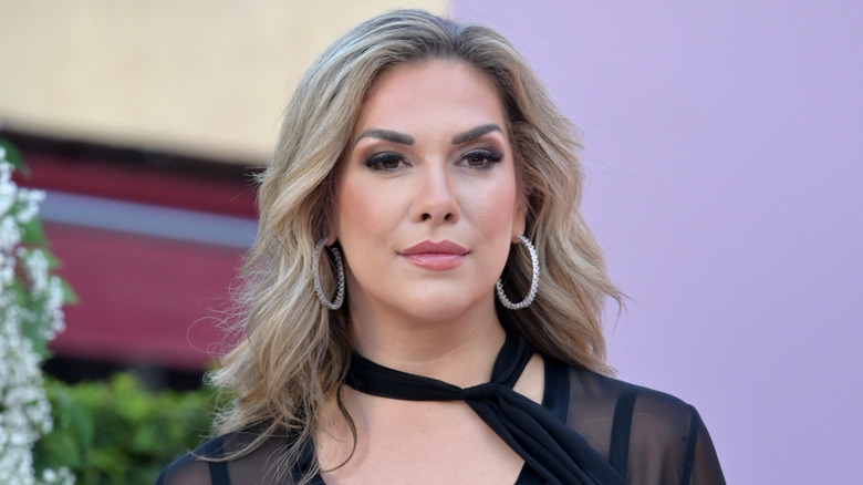 Allison Holker wearing a black see through top with a black neck tie and silver hoop earrings