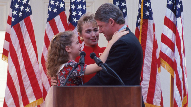 Chelsea Clinton embracing Bill Clinton at podium surrounded by American flags as Hillary Clinton smiles in between them