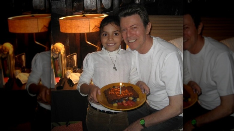 Young Lexi Jones holds a birthday cake and stands beside a seated David Bowie.