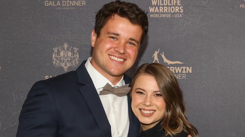 Chandler Powell wearing a blue tuxedo and grey bowtie with his arm around Bindi Irwin wearing a black collared dress