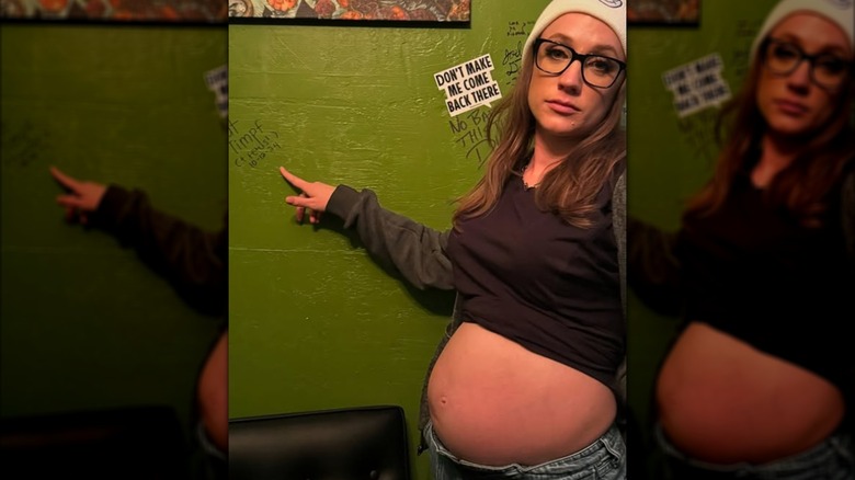 A pregnant Kat Timpf points at her signature on a wall.