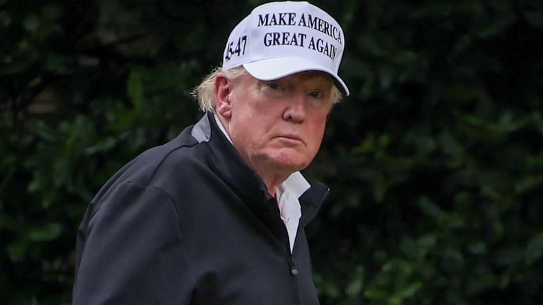 Donald Trump outside, wearing a MAGA hat, face red, circles under eyes, looking at camera