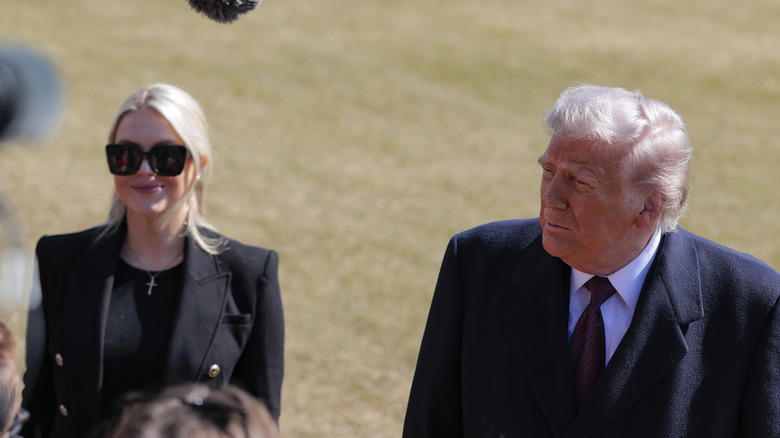 Karoline Leavitt smiling while wearing sunglasses standing next to Donald Trump during a press gaggle