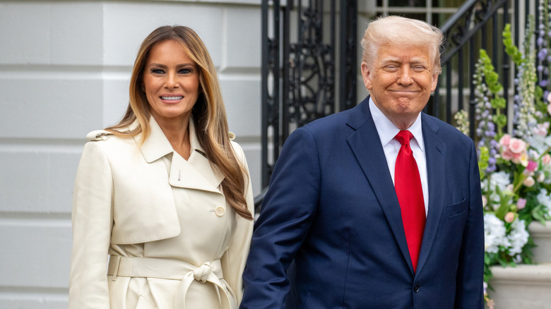 Melania Trump wearing a trench coat and Donald Trump wearing a suit outdoors as they both smile