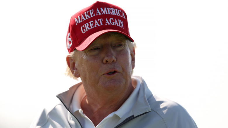 Donald Trump wearing his red MAGA hat on an overcast golf course.