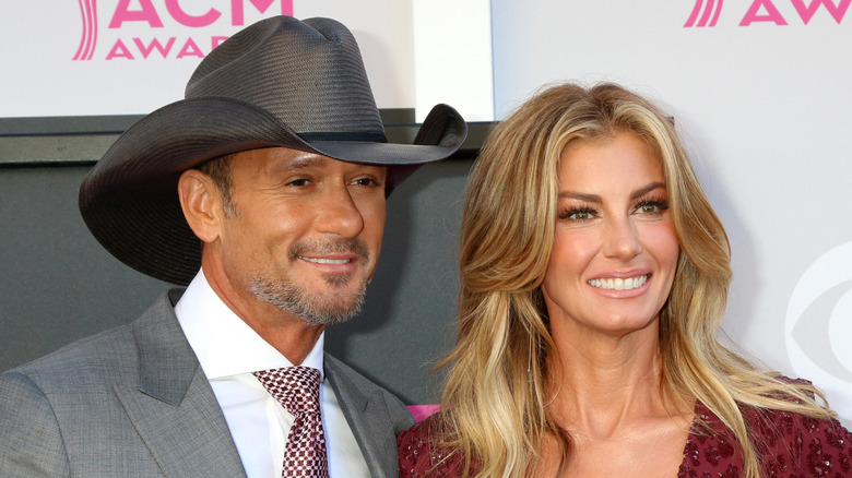 Tim McGraw wearing a grey suit and patterned tie with a black cowboy hat with Faith Hill wearing a red sequined long sleeved gown