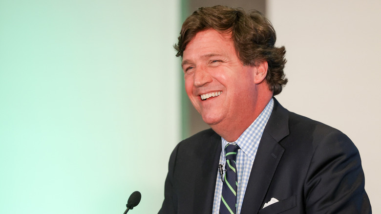 Tucker Carlson laughing in front of a microphone while wearing a grey suit in 2023
