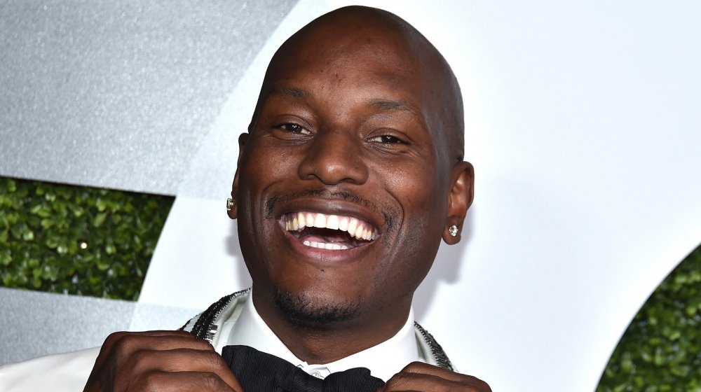 Tyrese Gibson in a white suit, smiling big as he adjusts his black bow-tie on the red carpet