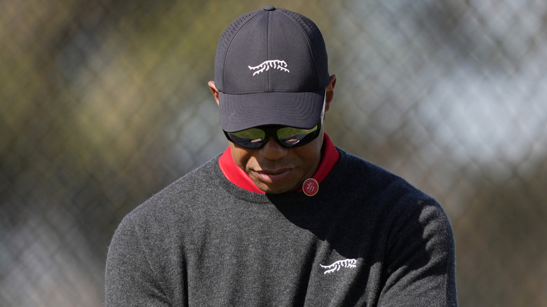Tiger Woods looking towards the ground while golfing in a dark grey ensemble with a matching hat
