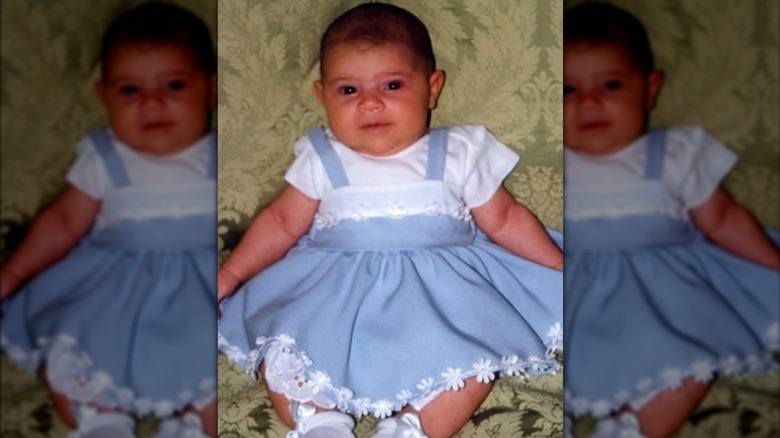 Vanna White's daughter, Gigi" Santo Pietro, wearing a blue pinafore and sitting on a couch as an infant.