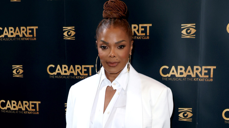 Janet Jackson wearing a white collared shirt, white blazer and large gold hoop earrings with her hair braided and in a bun