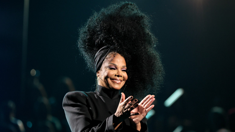 Janet Jackson clapping and smiling wearing a black long-sleeved gown and black headwrap with a large afro hairstyle