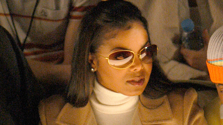 Janet Jackson looking to the side while wearing tinted sunglasses, a white turtleneck, and tan jacket