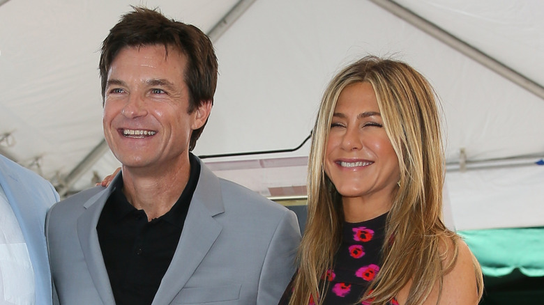 Jason Bateman wearing a grey suit jacket and black shirt with Jennifer Aniston wearing a high collared black and floral dress while smiling