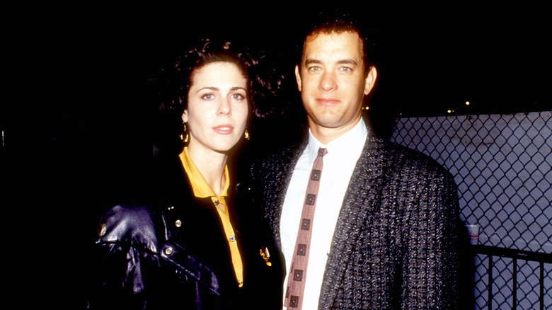 Young Rita Wilson and Tom Hanks posing in front of chain-link fence