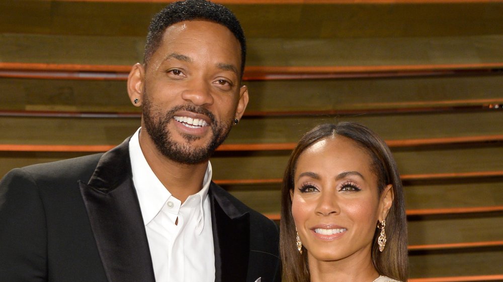 Will Smith in a black blazer and white shirt with no tie, Jada Pinkett Smith in a white-and-gold dress, both smiling