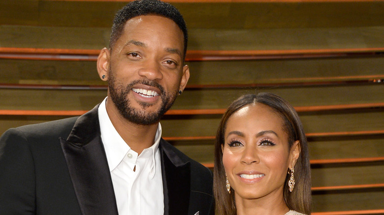 Will Smith in a black blazer and white shirt with no tie, Jada Pinkett Smith in a white-and-gold dress, both smiling