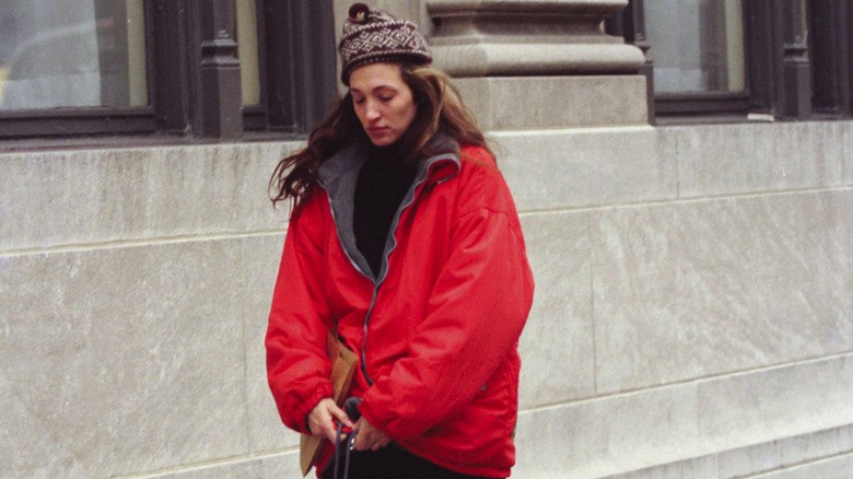 Wearing a red jacket and a beanie hat, Carolyn Bessette-Kennedy looks toward the ground.