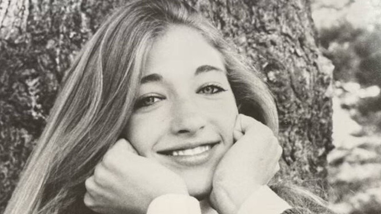 A young Carolyn Bessette-Kennedy flashes a wide smile, her hands resting on her cheeks.