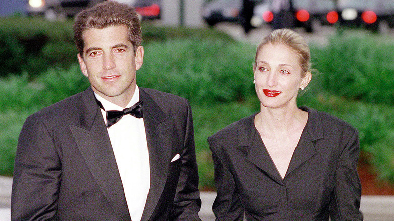 John F. Kennedy Jr. wearing a tuxedo outdoors with Carolyn Bessette-Kennedy, who is wearing a black tailored jacket and red lipstick.