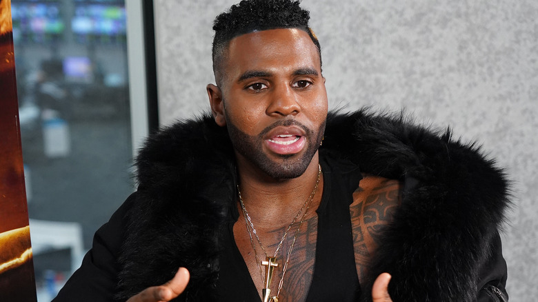 Jason Derulo, looking confused, explaining something, wearing a fur coat and black tank top