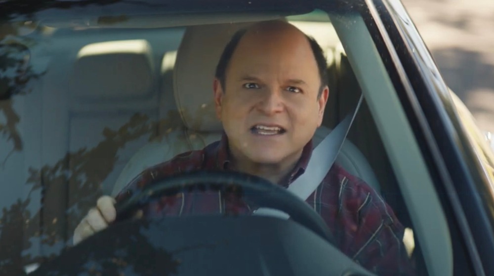 Jason Alexander looking straight ahead from the driver's seat of a car
