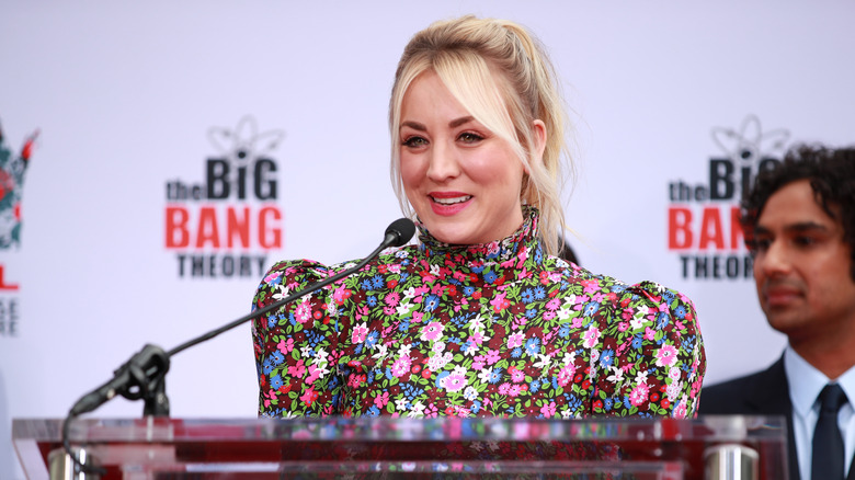 Kaley Cuoco wearing a high collared long sleeved floral dress speaking into a microphone on a podium