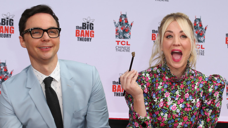Jim Parsons wearing.a light blue suit, black tie and glasses with Kaley Cuoco wearing a high collared long sleeved floral dress