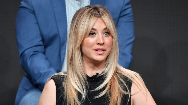 Kaley Cuoco with long blonde hair wearing a black sleeveless dress speaking on a panel for "Harley Quinn"