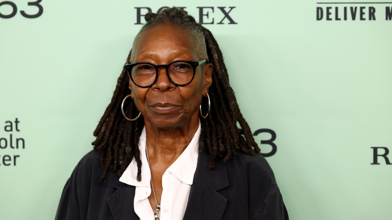 Whoopi Goldberg smiling for a picture while wearing a black blazer and white, open-collared shirt with silver hoop earrings