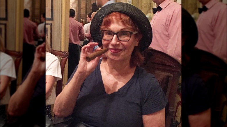 Joy Behar wearing glasses, a hat, and no makeup while holding a cigar to her mouth