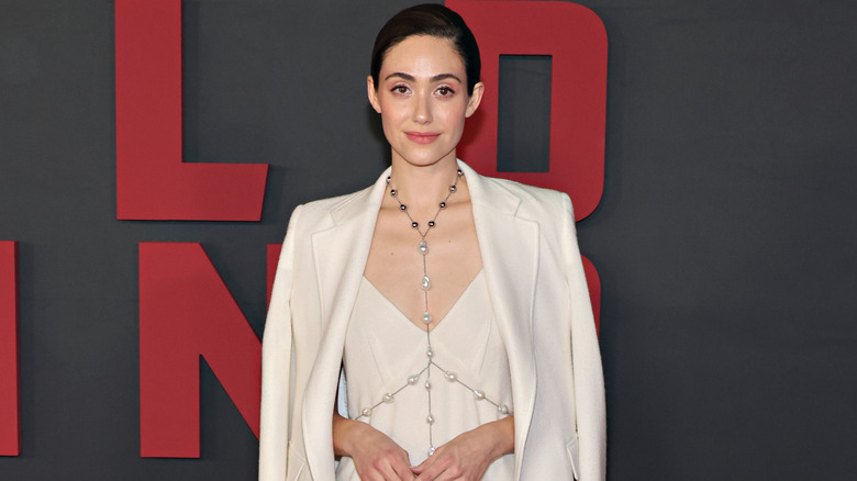 Emmy Rossum posing in front of black background with red lettering in 2023