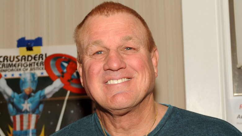 Reb Brown smiling at a fan expo in 2014 with a Captain America poster behind him.