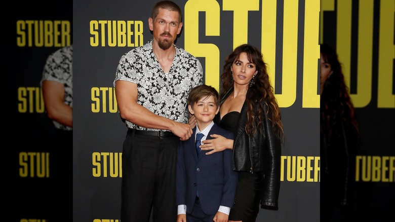 Steve Howey and Sarah Shani rest their hands on their son as they gaze away from the camera.