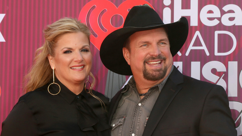 Trisha Yearwood and Garth Brooks smile on the red carpet while Yearwood wears a black blouse and Brooks wears a black cowboy hat and matching blazer