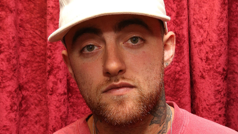 Mac Miller in a white hat standing in front of a red backdrop