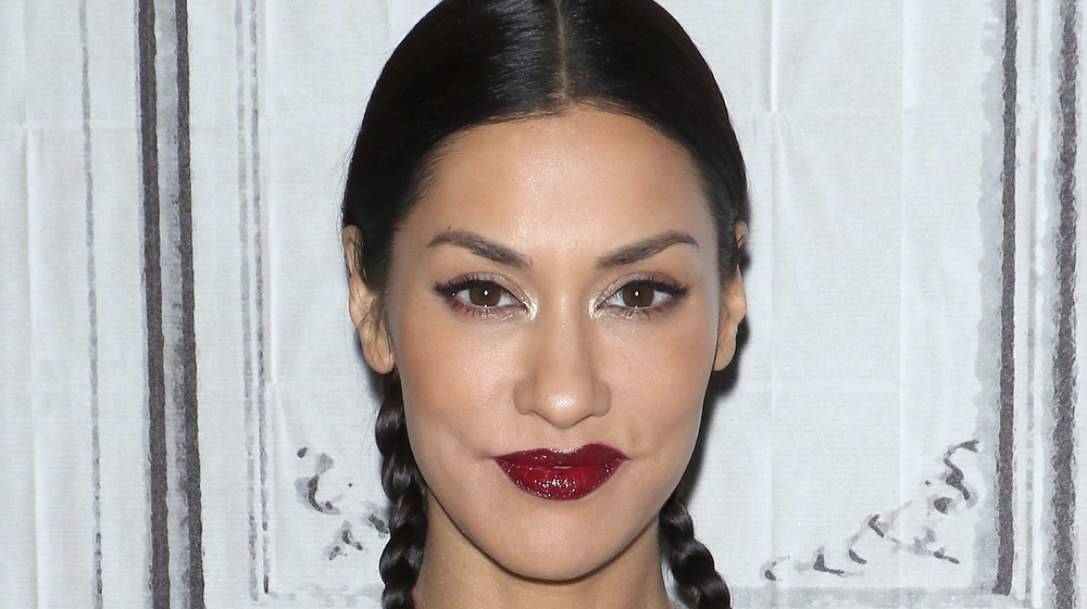 Janina Gavankar wearing dark red lipstick and braids smiling