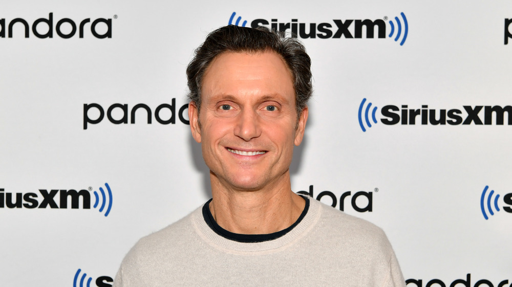 Tony Goldwyn smiles in front of a camera wearing a cream sweater