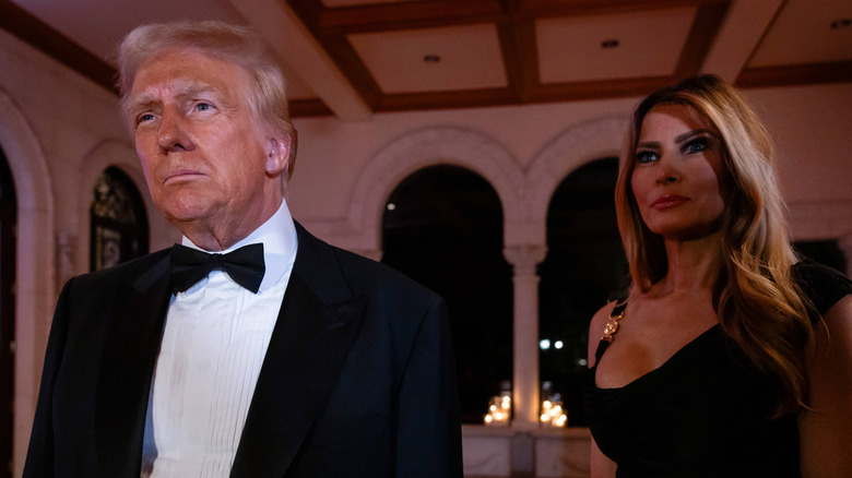 Donald Trump staring ahead while Melania Trump stands behind him smiling