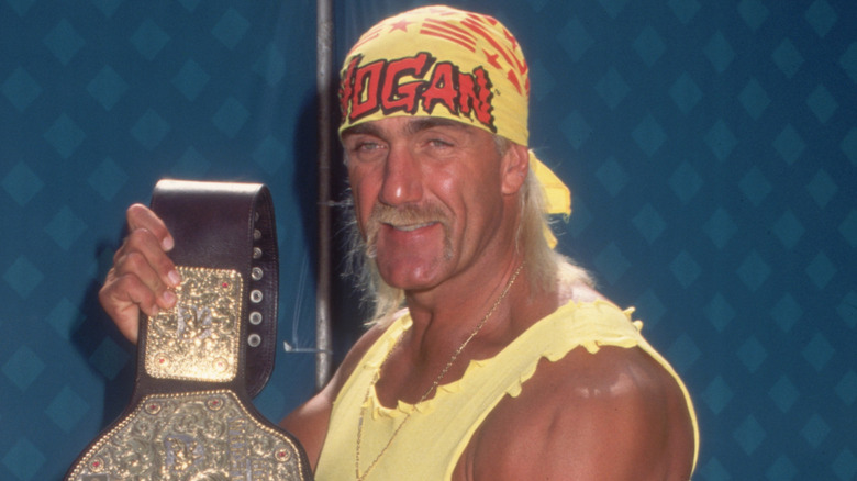 American wrestler Hulk Hogan displaying his championship belt. Hogan wears a costume consisting of a yellow 'HOGAN' bandana, a yellow muscle top, a weightlifter's belt, red spandex pants, and yellow cowboy boots.